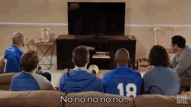 a group of people are sitting on a couch watching a television and one of them is wearing a number 18 jersey
