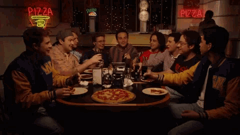 a group of people are sitting at a table with pizza in front of a pizza sign
