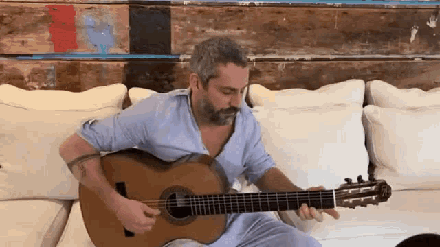 a man with a beard is sitting on a couch playing an acoustic guitar .