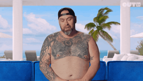 a man with tattoos on his chest is sitting on a blue couch in front of a palm tree