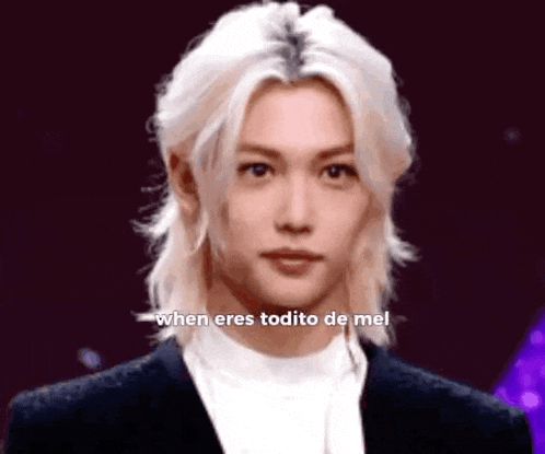 a young man with long blonde hair is wearing a black suit and a white turtleneck .