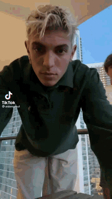 a young man with blonde hair is standing on a balcony with a city in the background .