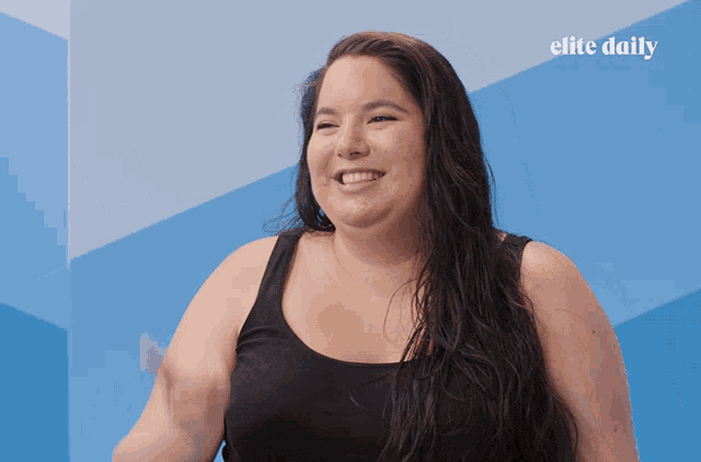 a woman in a black tank top is smiling in front of a blue wall with elite daily written on it