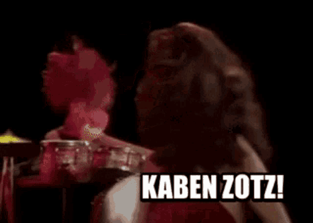 a woman in a red dress is dancing in front of a drum set and says " suuuu-kal-da-tu "