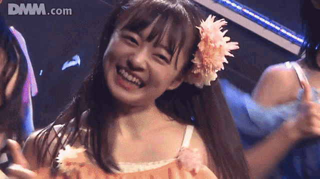 a girl with a flower in her hair smiles in front of a dmm.com logo