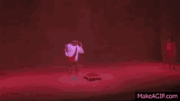 a man in a white shirt and red tie is dancing on a stage with red lights behind him and the words makeagif.com below him