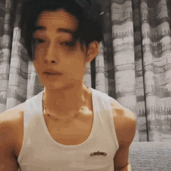 a man wearing a white tank top and a necklace is standing in front of a curtain