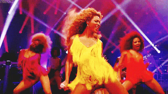 a woman in a yellow dress is singing into a microphone while dancing on a stage with other dancers .