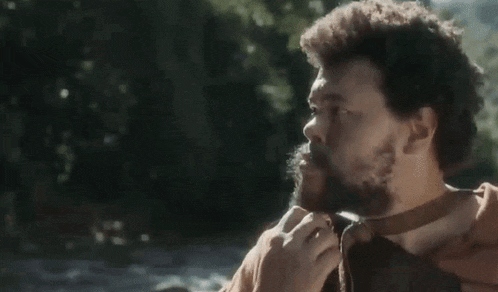a man with a beard is tying a belt around his neck while standing next to a river .