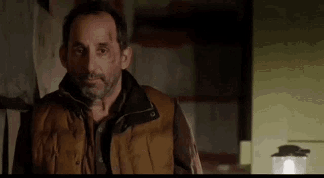 a man in a brown vest is standing in a dark room with a lantern in the background .