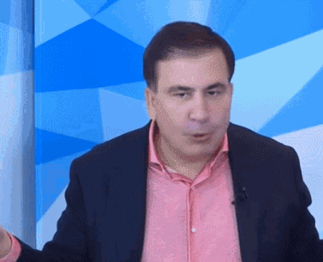 a man wearing a pink shirt and a black jacket is standing in front of a blue background