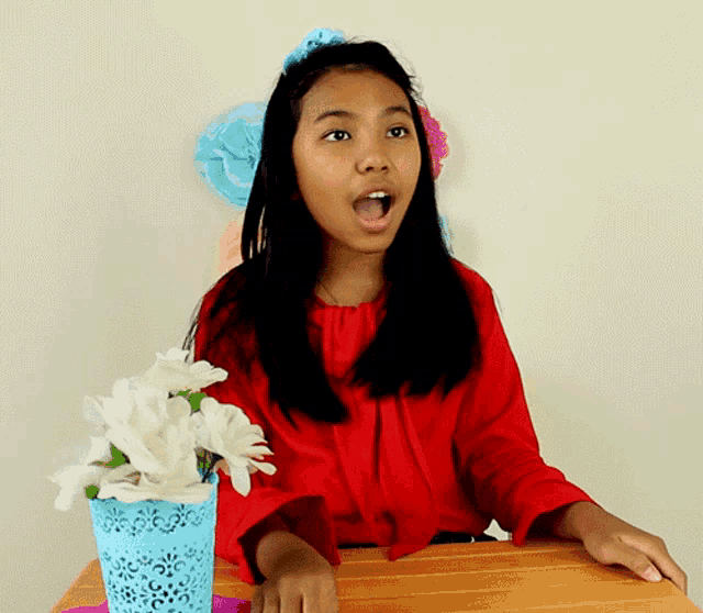 a girl in a red shirt is sitting at a table with flowers in a blue basket