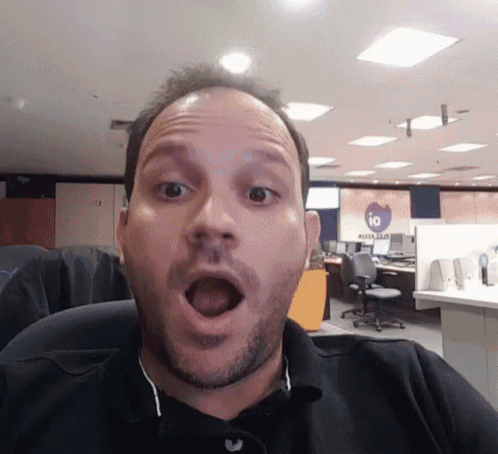 a man with a surprised look on his face in an office with a sign that says io in the background