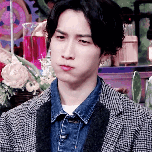 a young man wearing a plaid jacket and a denim shirt looks at the camera
