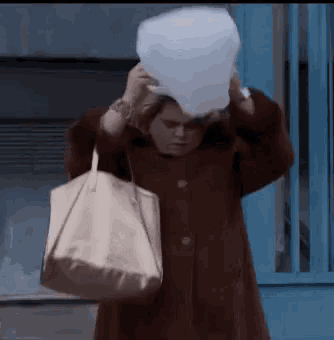 a woman wearing a brown coat is carrying a white bag and a bucket on her head