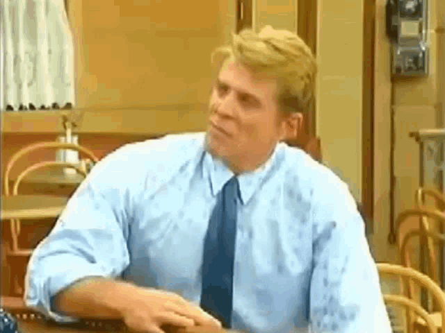 a man in a blue shirt and tie is sitting at a table in a kitchen .