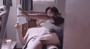 a man is laying on a bed reading a book while another man sits on his lap .
