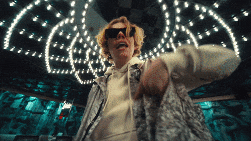 a man wearing sunglasses and a white hoodie stands in front of a chandelier