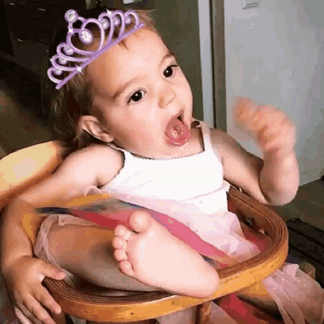 a baby girl wearing a tiara is sitting in a high chair with her tongue out