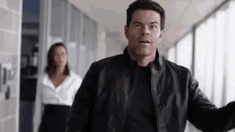 a man in a leather jacket is walking down a hallway with a woman in a white shirt behind him .