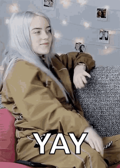 billie eilish is sitting on a couch and making a funny face while holding a cell phone .