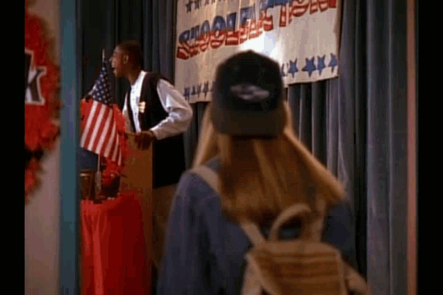 a man is giving a speech at a podium in front of an american flag while a woman looks on .