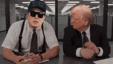 a man wearing a blue hat that says " make america great again " sits at a table with a man in a suit