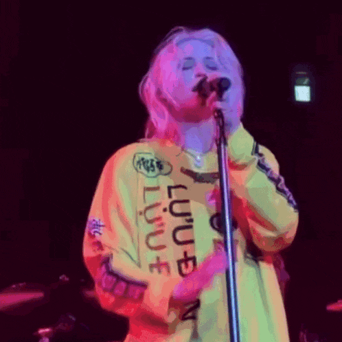 a woman is singing into a microphone while wearing a yellow sweatshirt that says ' lulu ' on it