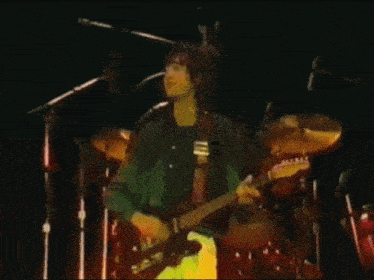 a man playing a guitar on stage with a drum set in the background