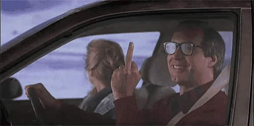 a man in glasses is driving a car and giving the middle finger .