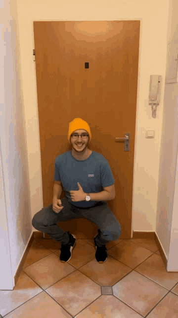 a man wearing a blue shirt and a yellow hat squats in front of a door and gives a thumbs up