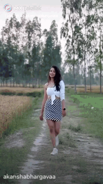 a woman in a white top and black and white skirt is walking on a dirt road