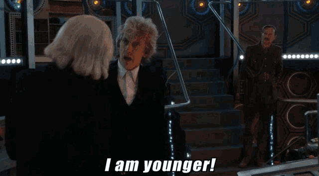 two men are standing next to each other in a room and one of them says i am younger .