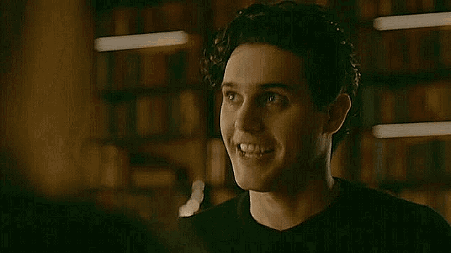 a man in a black shirt is standing in front of a bookshelf and talking to another man .