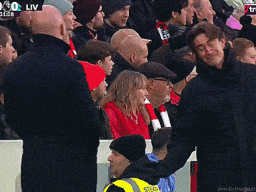 a man in a black jacket stands in front of a crowd with a steward in a yellow vest behind him