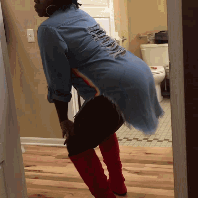 a woman wearing a denim shirt and red boots stands in front of a toilet