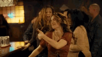 a group of people are standing around a bar and one woman is pointing at something .