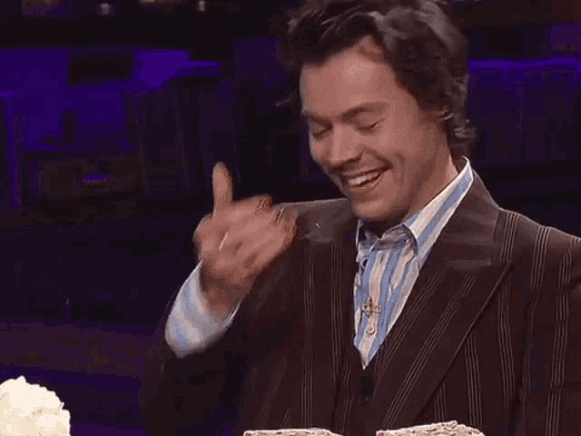 harry styles is wearing a suit and giving a thumbs up while standing in front of a cake .