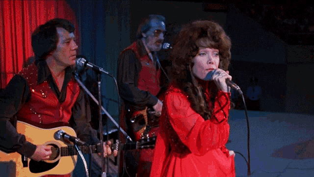 a woman in a red dress is singing into a microphone while two men play guitars