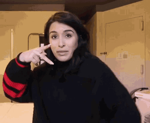a woman wearing a black sweater with red stripes on the sleeves points at her face