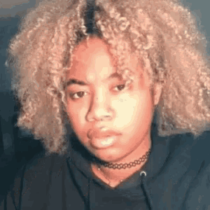 a young woman with blonde curly hair is wearing a choker necklace and a black hoodie .