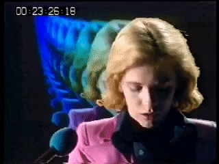 a woman in a pink jacket is talking into a microphone with the time of 23:26:18 on the screen behind her