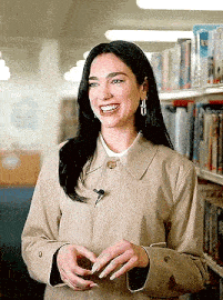 a woman in a trench coat is standing in front of a bookshelf in a library and smiling .