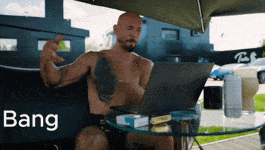a shirtless man sits at a table with a laptop and bang written on the screen