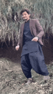a man in a brown jacket and black pants is standing in a field of tall grass