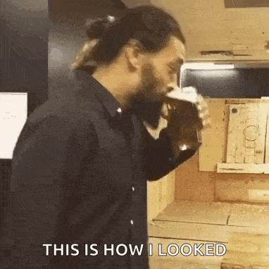 a man in a black shirt is drinking from a cup while standing in a kitchen .