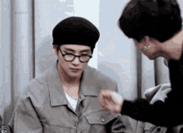 two men are talking to each other in a room . one of the men is wearing glasses and a beret .