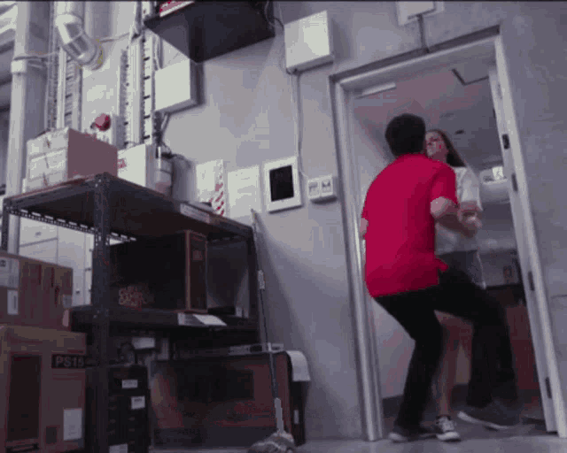 a man in a red shirt is standing in a room with boxes on the floor and a ps15 box on a shelf