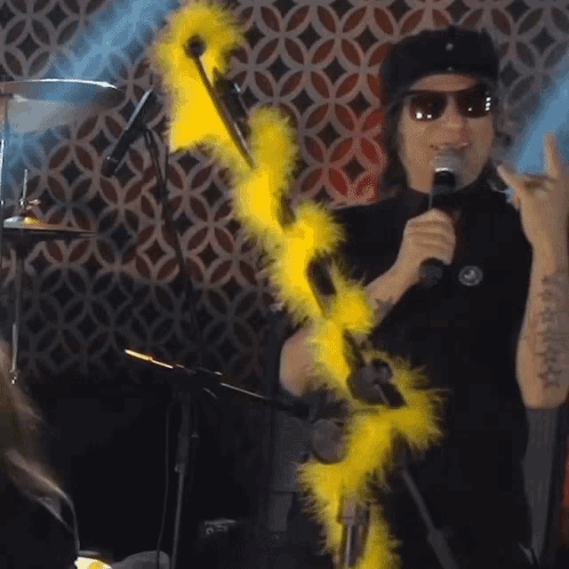 a man wearing sunglasses is singing into a microphone while holding a yellow boa around his neck
