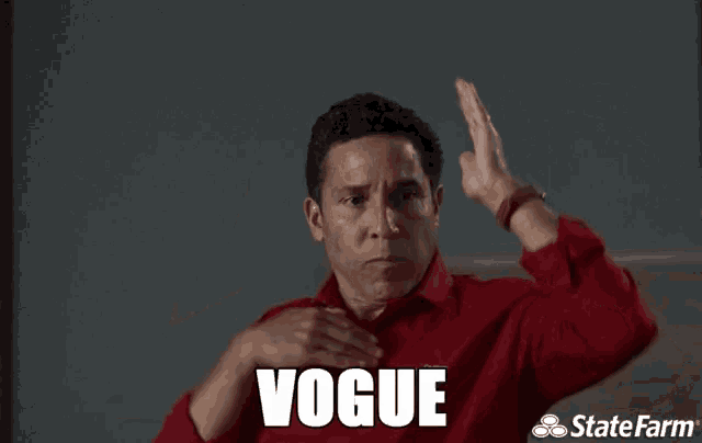 a man in a red shirt is making a gesture with his hands and the word vogue is on the screen behind him .